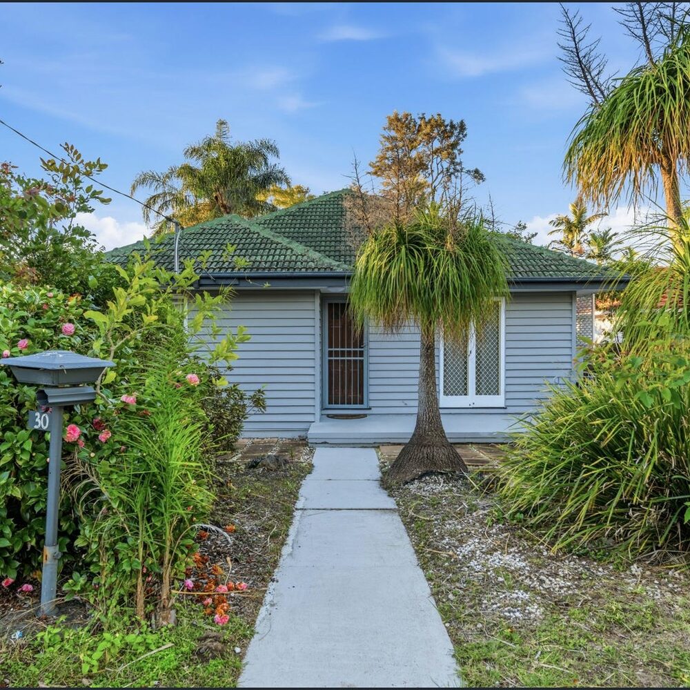 Blue weatherboard cottage behind a garden path and mature planting, Wavell Heights